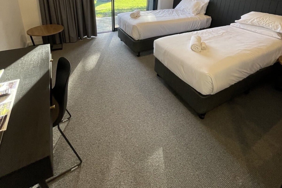 Une chambre d’hôtel avec deux lits, un bureau et une vue sur un paysage verdoyant, visible à travers une porte vitrée.