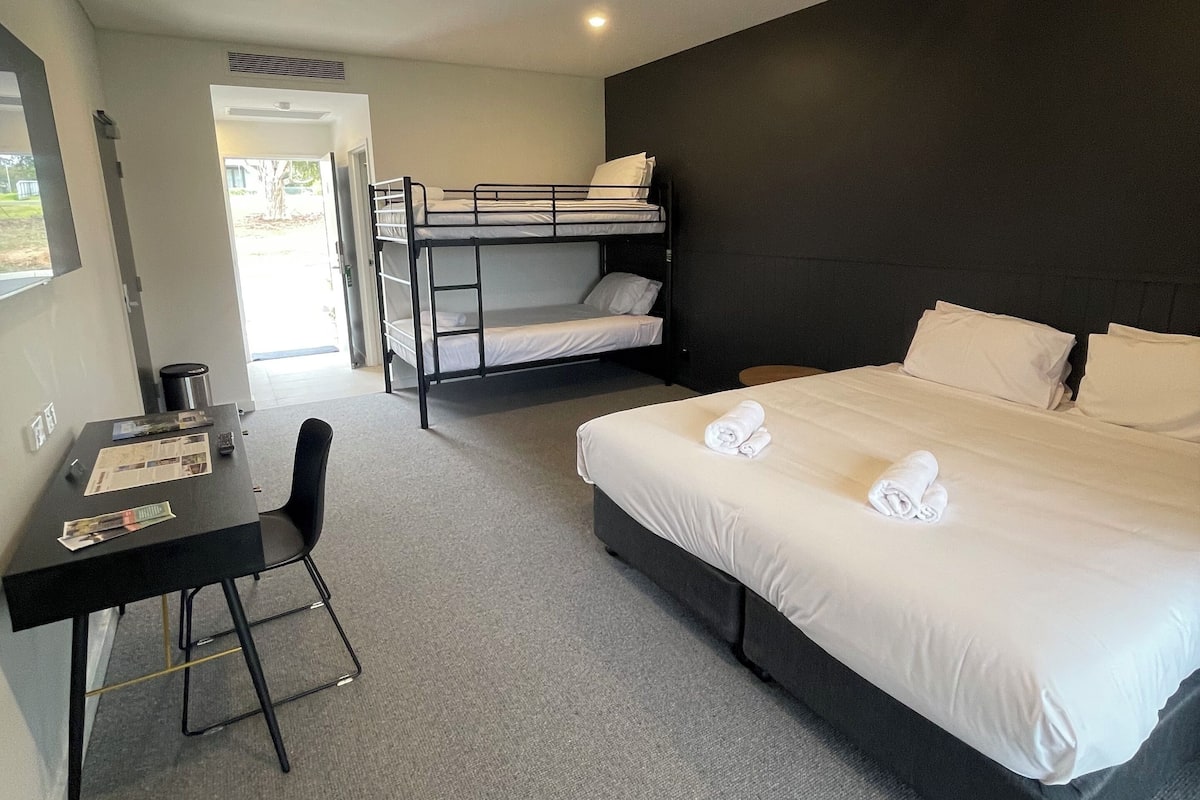 Une chambre d’hôtel avec un lit superposé, un bureau avec une chaise et un grand lit avec des serviettes soigneusement pliées.