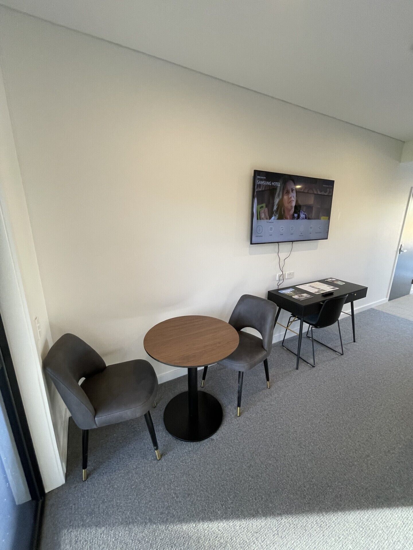Une petite pièce moderne avec une table ronde et deux chaises, un téléviseur à écran plat fixé au mur et un sol recouvert de moquette.