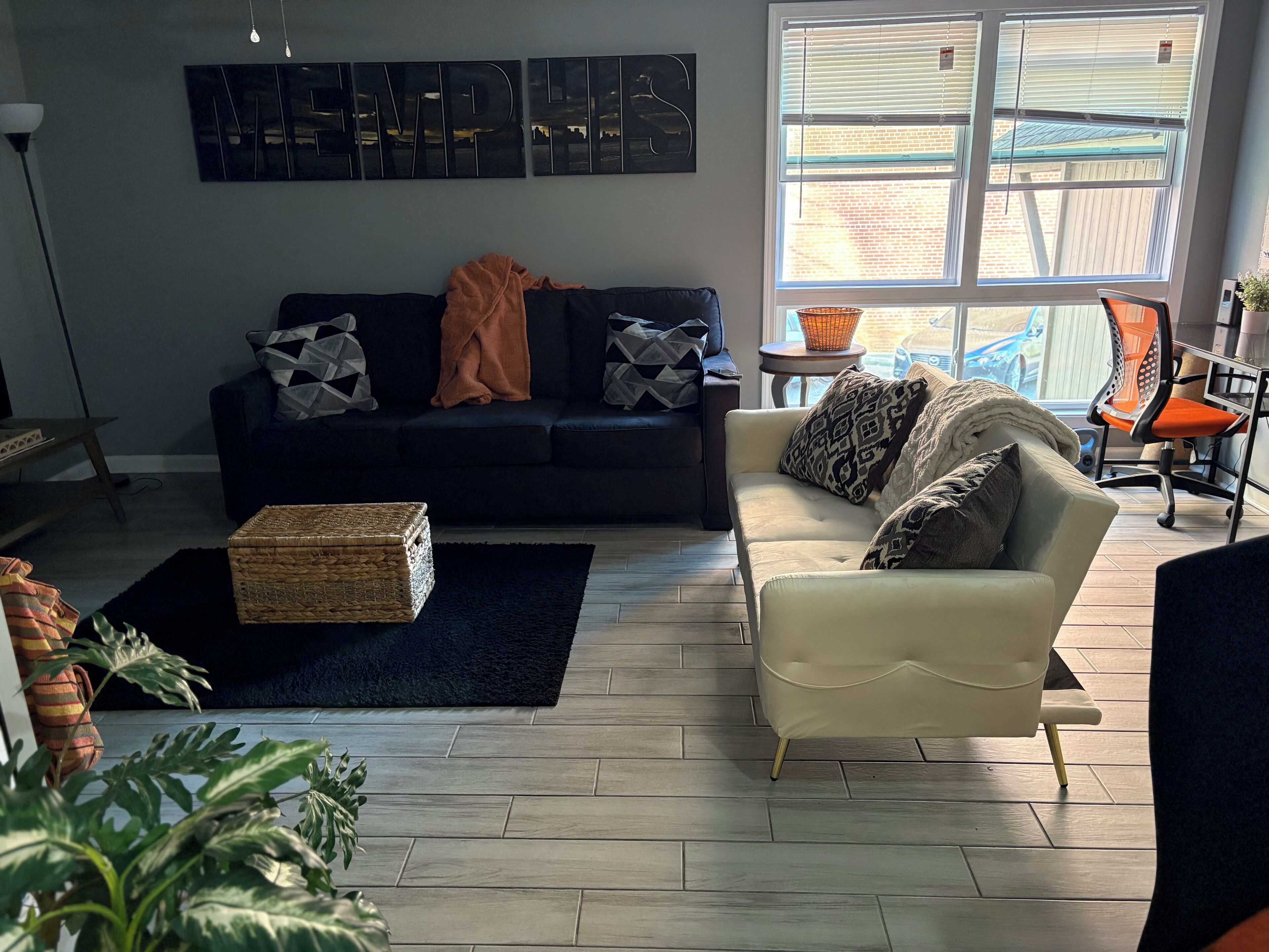 A living room with a dark-colored sofa, a white armchair, and a small side table. There is a large window with blinds, a floor lamp, and a decorative wall piece.
