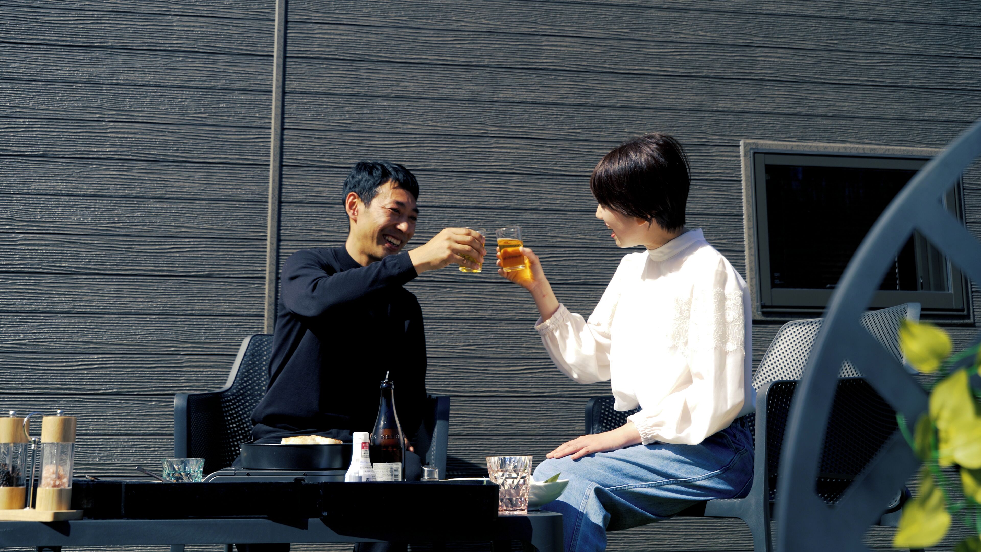 Deux personnes trinquent avec des boissons à une table en plein air, où l’on aperçoit une chaise en métal, une bouteille et un verre.