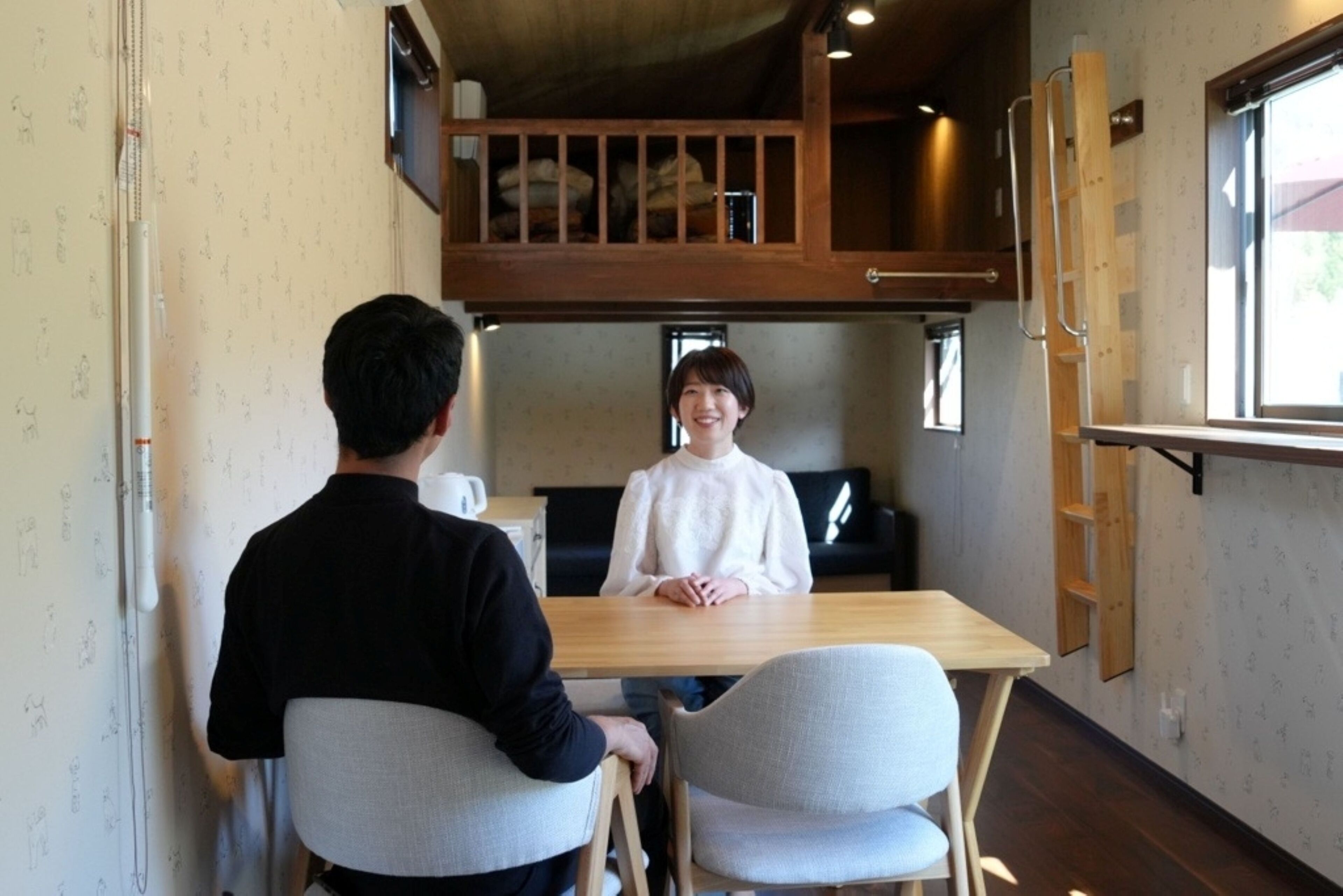Una habitación con una mesa de madera, dos sillas, una persona sentada a la mesa y otra de pie frente a ella.