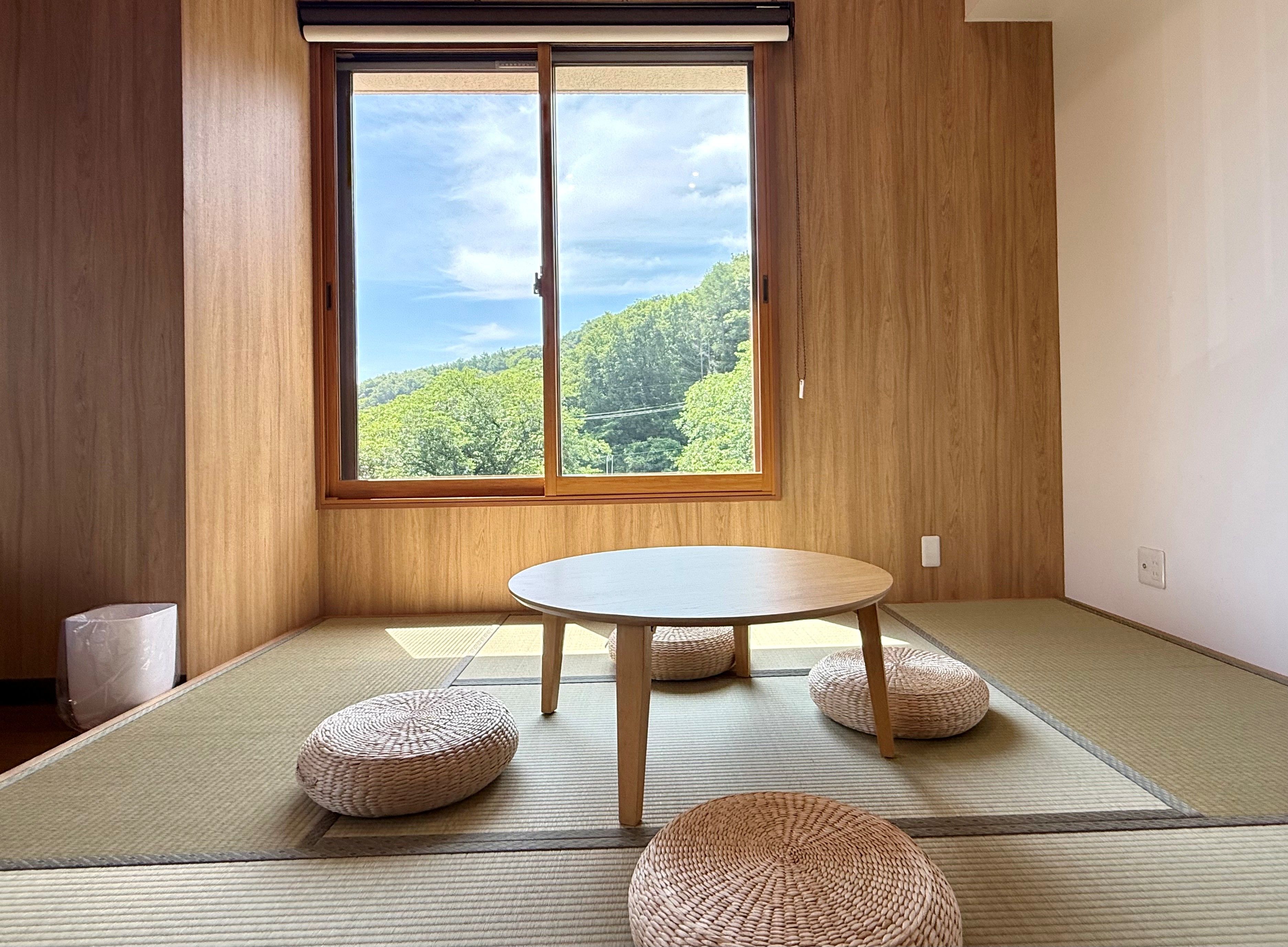 Ein Zimmer mit Tatami-Matten, einem runden Tisch und einem Fenster mit Blick auf einen Wald.