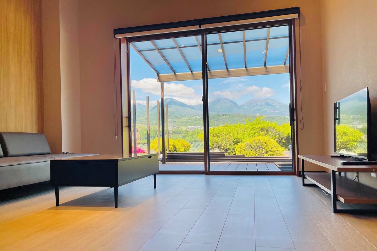 Un salon moderne comprenant un canapé, une table basse, un téléviseur et une grande porte-fenêtre coulissante donnant sur un balcon avec vue sur la montagne.
