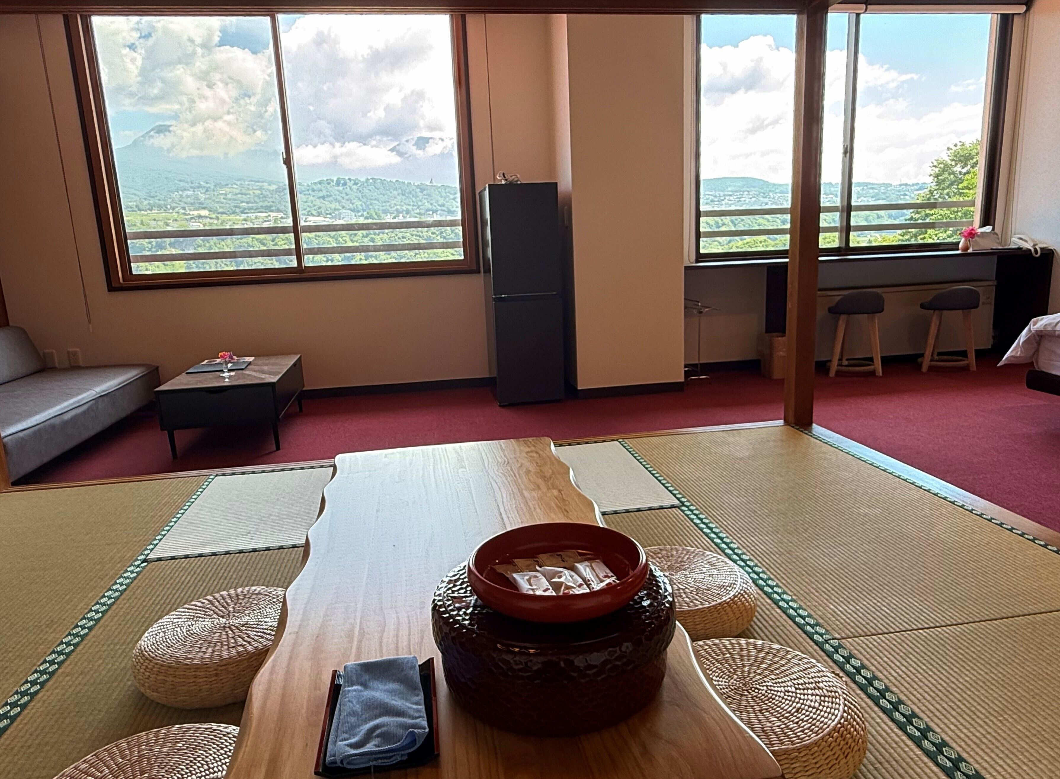 A room with tatami mats, a table with a ceramic pot, a sofa, a refrigerator, and a view of a green landscape through large windows.