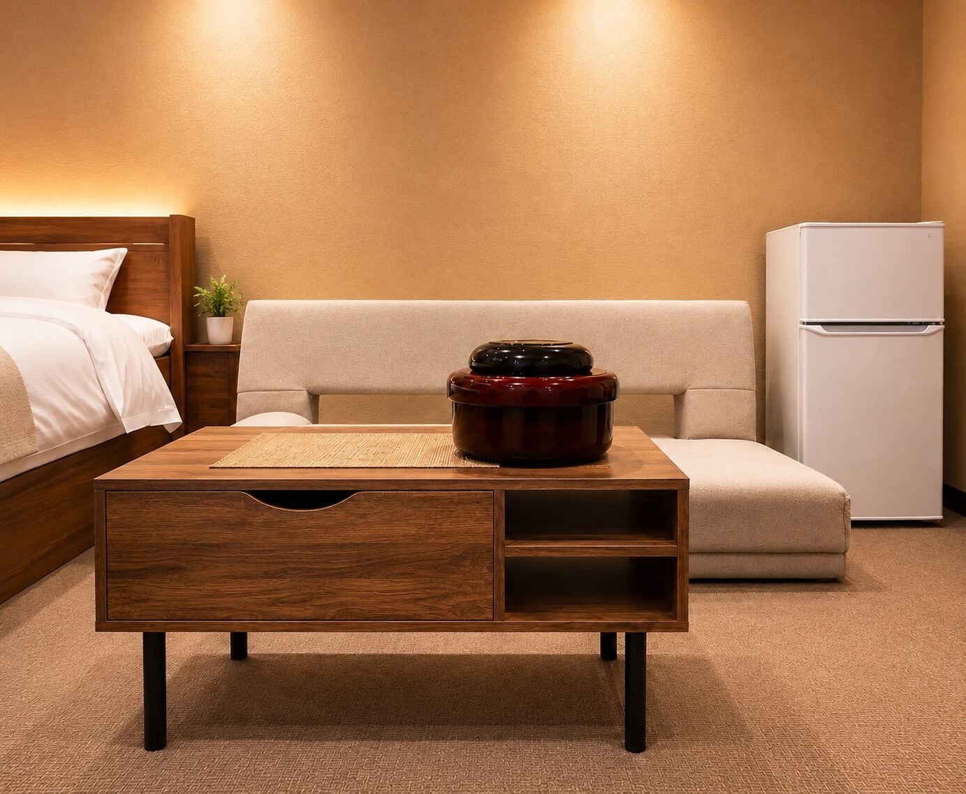 A hotel room with a wooden nightstand, a red pot, a white refrigerator, and a beige sofa.