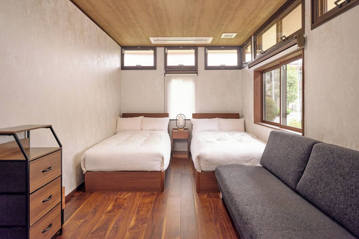 Une chambre avec deux lits, une commode en bois, un canapé gris et une fenêtre avec des stores.