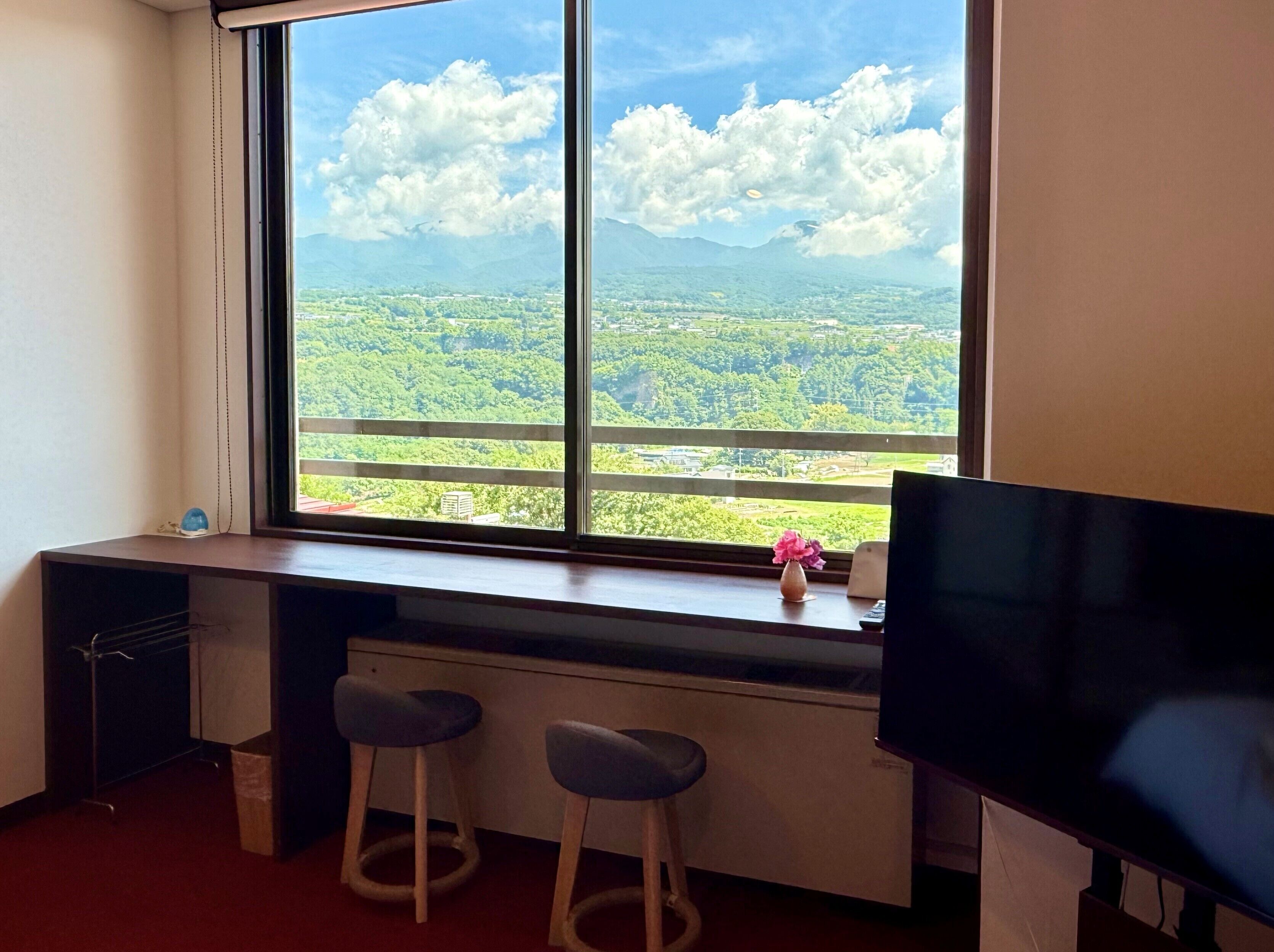 A room with a large window, a desk, two stools, and a television.