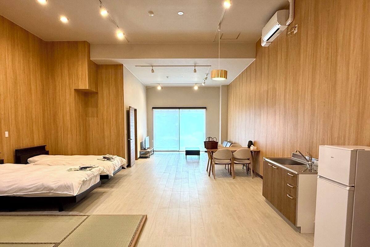 Une chambre d’hôtel moderne dotée d’un grand lit, d’un bureau avec des chaises, d’une kitchenette et d’une porte coulissante donnant sur un balcon.