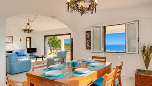 A dining area with a table set for four, a blue sofa, a TV, and a view of the sea. - Your perfect Algarve retreat with a pool (Sao Rafael)
