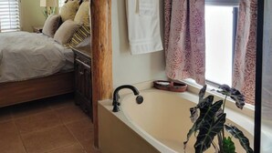 Un dormitori amb un llit gran, una columna de fusta, un bany amb banyera i una finestra amb cortines estampades.