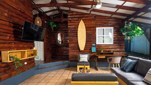 A cozy log cabin interior with a wooden TV, a surfboard, a sofa, and a dining area. - Charming 3-bedroom beach cabin with Starlink WiFi, AC in enchanting Tamanique (Tamanique)
