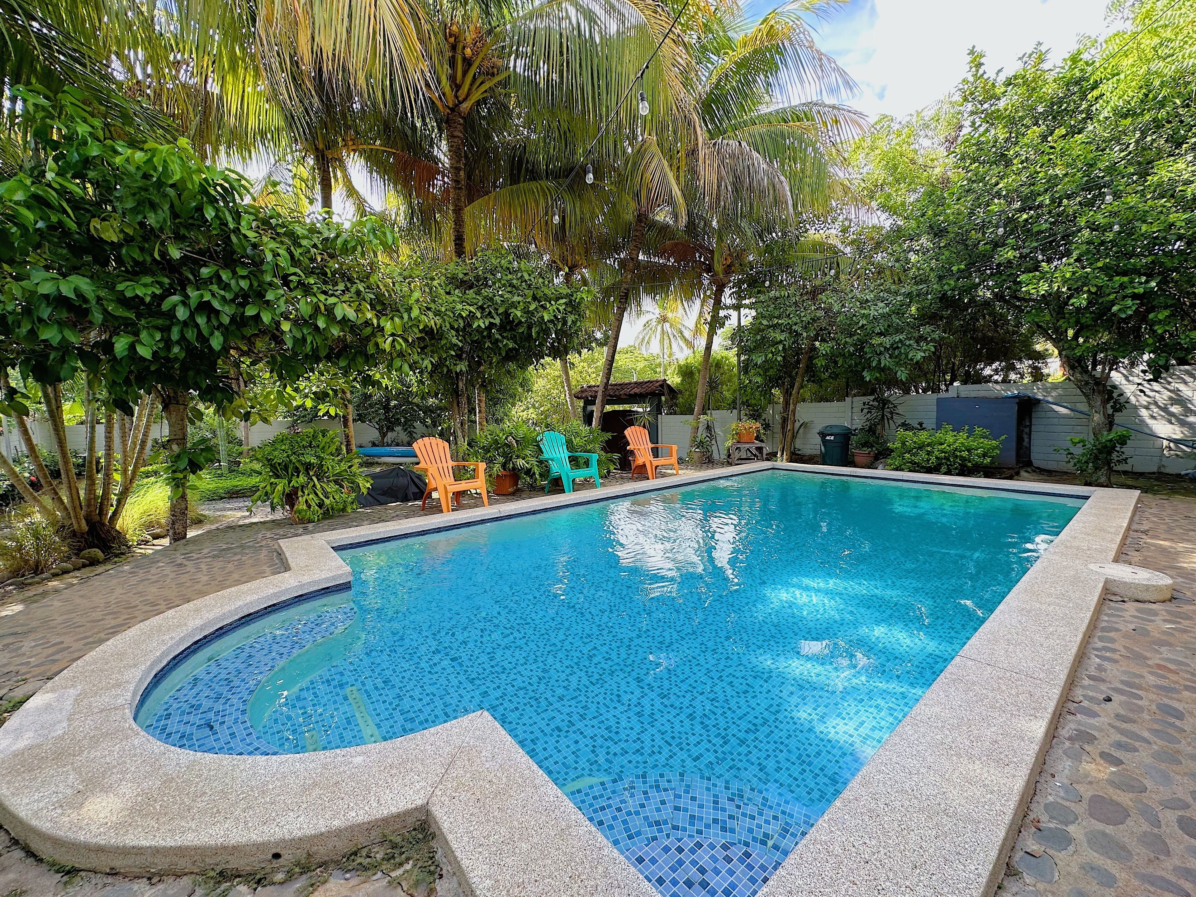 A rectangular swimming pool with a tiled edge, surrounded by greenery and three colorful chairs.