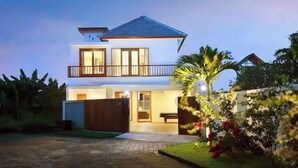 A two-story house with a balcony, a garage, and a well-maintained garden with flowers and a palm tree.