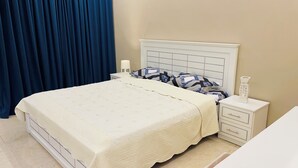 A neatly made bed with a white bedspread, a headboard with a grid pattern, and a bedside table with a lamp and a decorative item. - Amazing 3-bedroom apartment with WiFi in Ajman (Ajman)