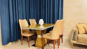 A dining area with a glass-top table and four chairs, a sofa with patterned cushions, and a blue curtain backdrop. - Amazing 3-bedroom apartment with WiFi in Ajman (Ajman)