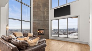 A spacious living room with a leather sofa, a fireplace, and large windows offering a mountain view.