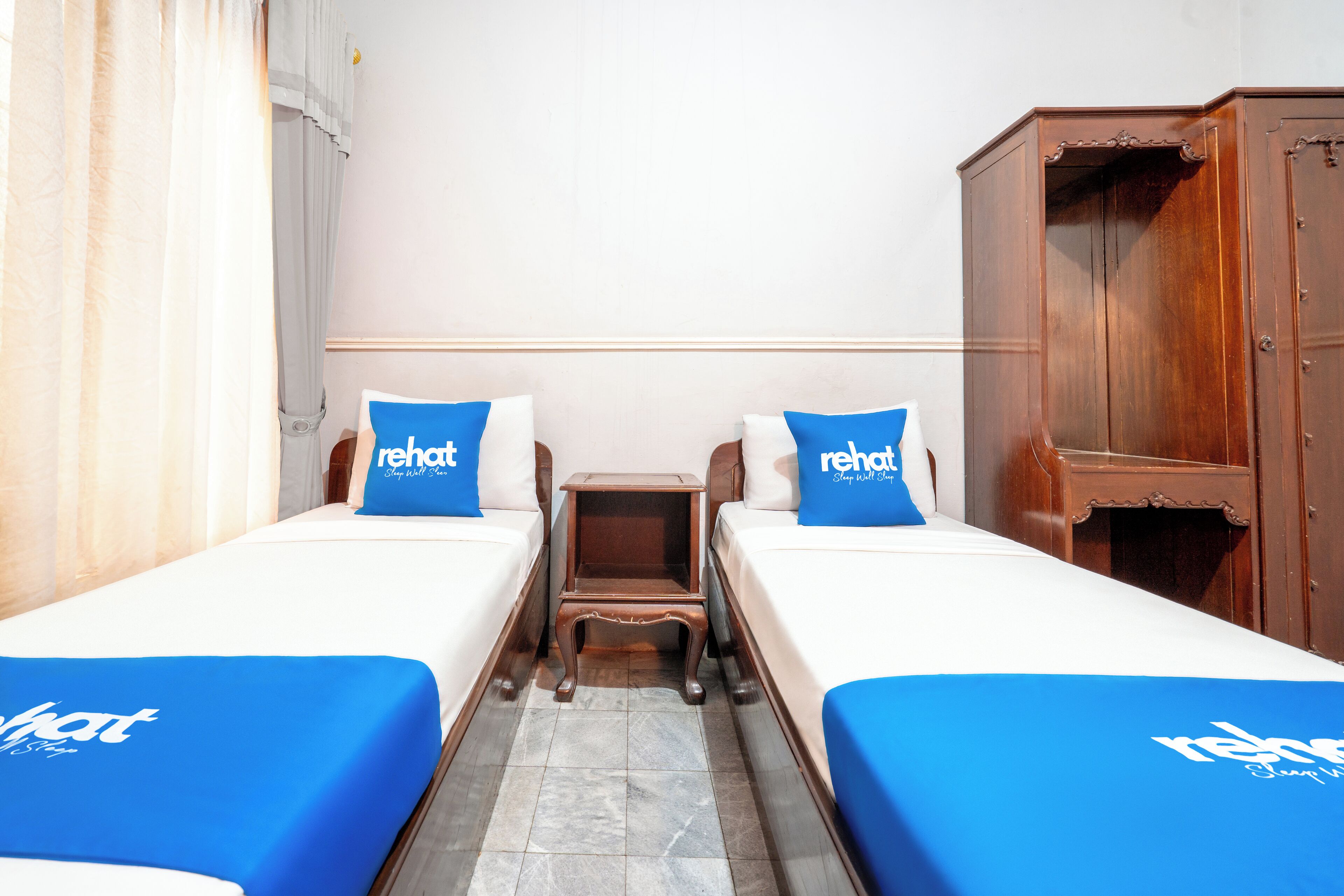 A hotel room with two single beds, each with a blue bedspread and matching pillows, a wooden nightstand, and a wooden wardrobe.