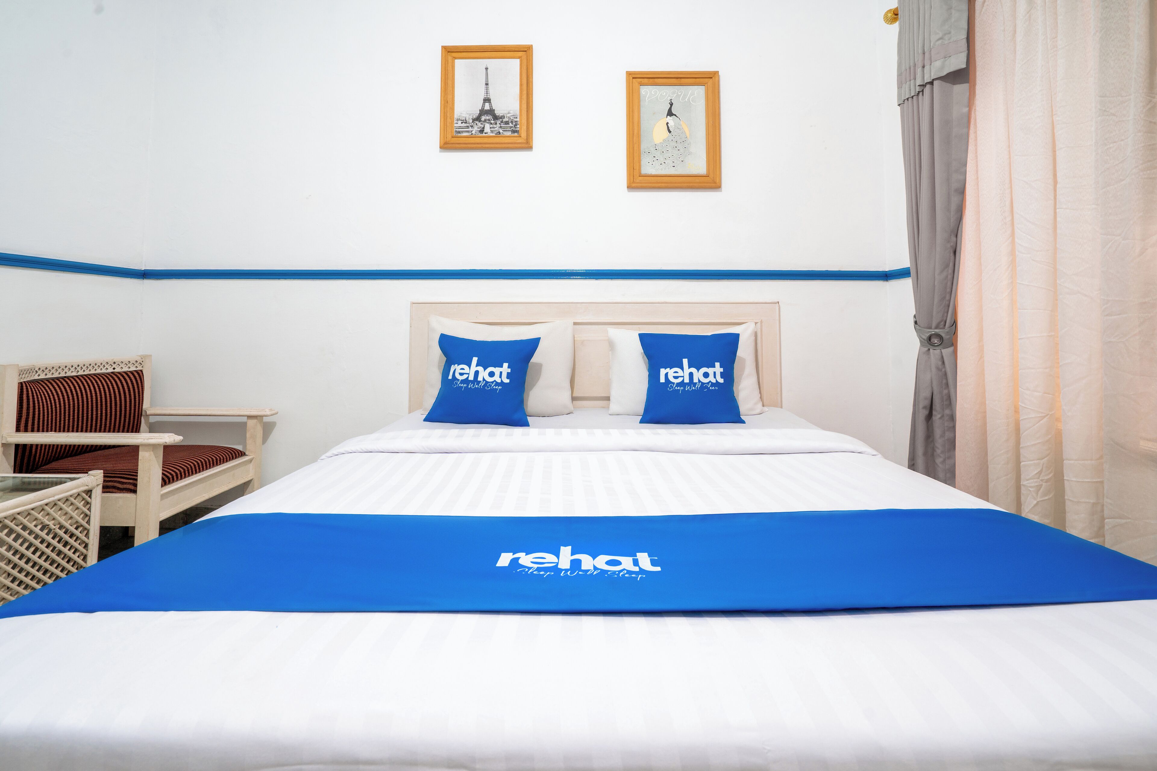 Habitación de hotel con cama, dos almohadas con la marca 'rehat', una silla y dos cuadros en la pared.