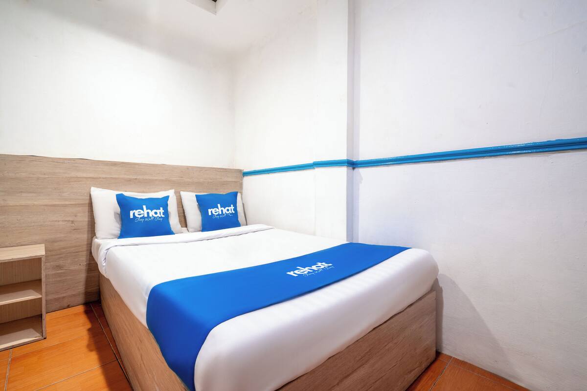 Ein Hotelzimmer mit hölzernem Kopfteil, einem Bett mit weißer Bettwäsche und blauen Akzenten, zwei Kissen mit 'rehat'-Branding und einem blauen Bettüberzug.