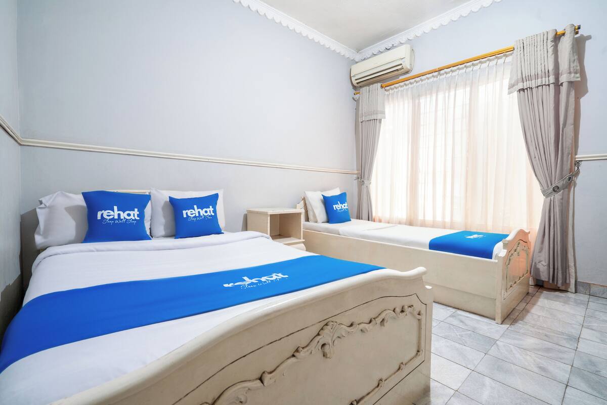 Ein Hotelzimmer mit zwei Betten, jeweils mit blauer Bettwäsche und Kissen, einem kleinen Nachttisch und einem Fenster mit Vorhängen.