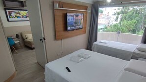 Habitación de hotel con dos camas, un televisor montado en un panel de madera, un balcón con vistas a edificios y árboles, y una pequeña zona de estar con una silla y una mesa.