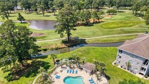 Un campo de golf con piscina y tumbonas, rodeado de árboles y un edificio.