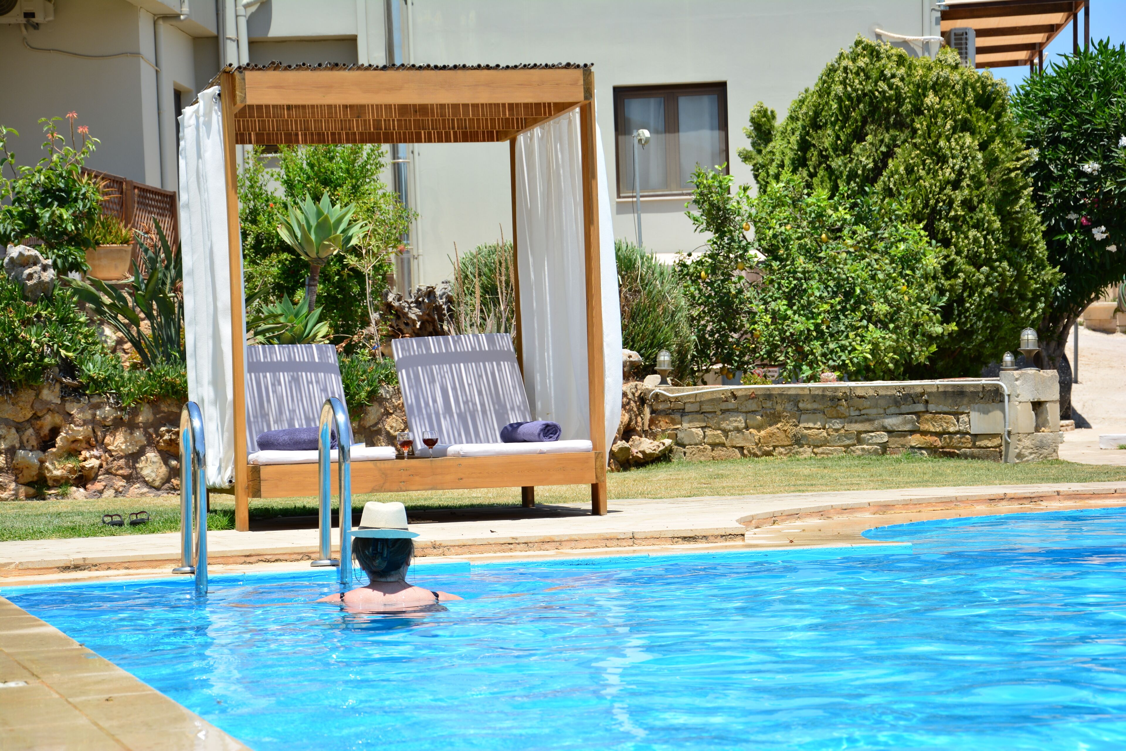 Ein Pool mit Holzterrasse und Sonnensegel, umgeben von Grünpflanzen und einer Steinmauer.