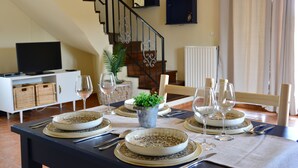 Mesa de comedor preparada para cuatro personas, con platos, vasos y cubiertos. Hay una planta en maceta sobre la mesa, y se aprecia una escalera al fondo.