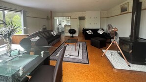 A modern living room with a sofa, coffee table, and a fireplace. There is a dining area with a glass table and chairs. The room has wooden flooring and a view of the kitchen in the background.