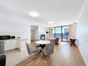 A modern apartment with a kitchen, dining area, and large windows offering a city view. - 7 Min to Train: Central Parramatta 2BR + Parking (Parramatta)