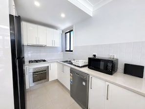 A modern kitchen with white cabinets, a stainless steel refrigerator, a microwave, an oven, and a dishwasher. - 7 Min to Train: Central Parramatta 2BR + Parking (Parramatta)
