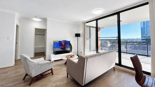 Ett modernt vardagsrum med en soffa, fåtölj och TV, stora fönster med utsikt över staden och en balkong.