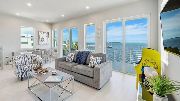 Ein modernes Wohnzimmer mit einem grauen Sofa, einem gemusterten Sessel und einem Couchtisch. Große Fenster bieten Blick auf das Meer und Gebäude.