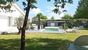 Area piscina moderna all'aperto con una sdraio, un piccolo spazio coperto con sedute e un edificio bianco sullo sfondo.