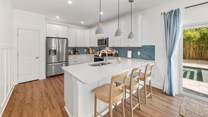 A modern kitchen with white cabinets, a stainless steel refrigerator, a large island with bar stools, pendant lighting, and a view of a swimming pool. - Coastal Chic, Heated Pool, Beachside, Custom Bunk Room (St. Augustine)