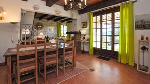 A rustic dining area with a wooden table and chairs, a stone wall, and a view through large windows with green curtains. - Awesome home in Saint-Laurent-du-Verdo (Saint-Laurent-du-Verdo)