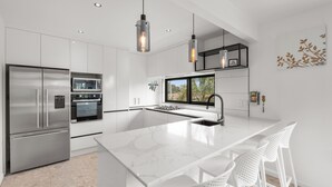 A modern kitchen with white cabinetry, stainless steel appliances, a marble island, and pendant lighting. - Orewa Beachcomber  (Orewa)