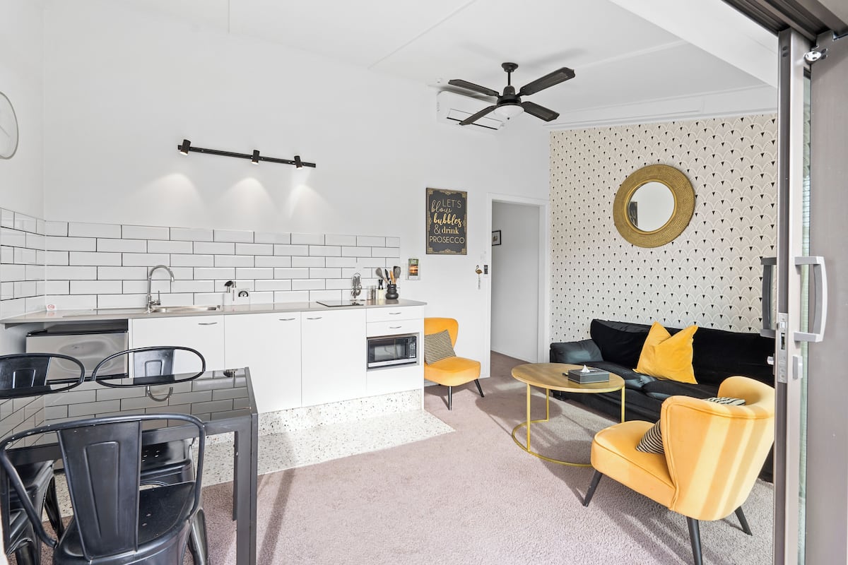 Eine moderne Küche mit weißen Schränken, einer schwarzen Arbeitsplatte und einem eingebauten Backofen. Ein Essbereich mit einem schwarzen Metallstuhl und einem gelben Sessel ist zu sehen. Ein schwarzer Deckenventilator ist sichtbar, und ein runder Spiegel hängt an der Wand.