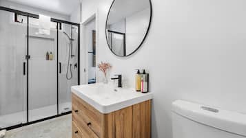 Une salle de bain moderne avec une douche à l’italienne en verre, un meuble-lavabo en bois avec un vasque blanche et un miroir rond au mur.