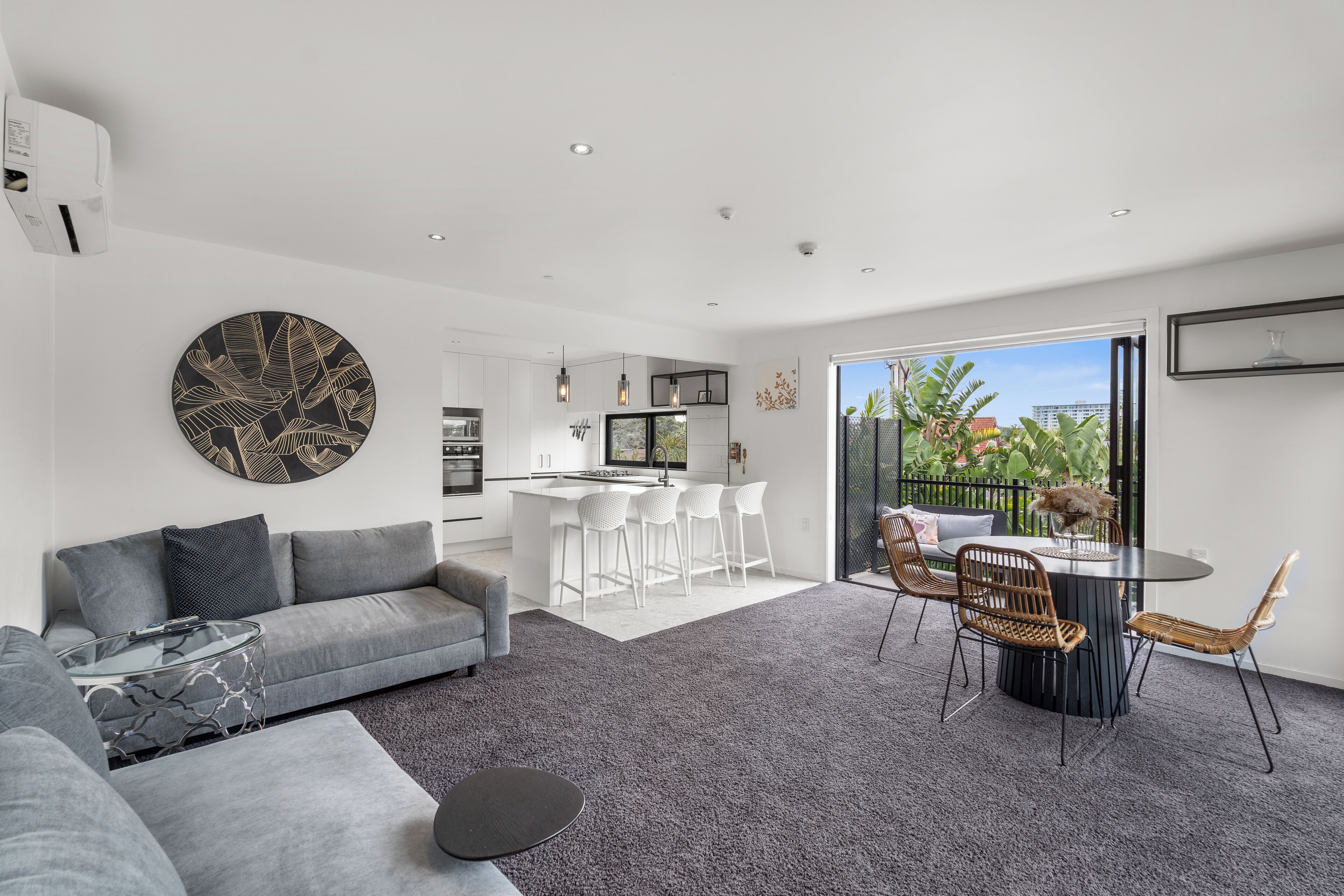 A modern living room with a grey sofa, a glass coffee table, and a dining area with a round table and wicker chairs.