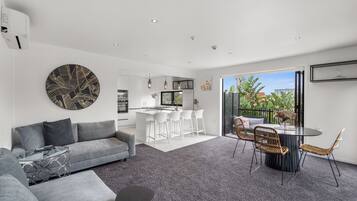 A modern living room with a grey sofa, a glass coffee table, and a dining area with a round table and wicker chairs.
