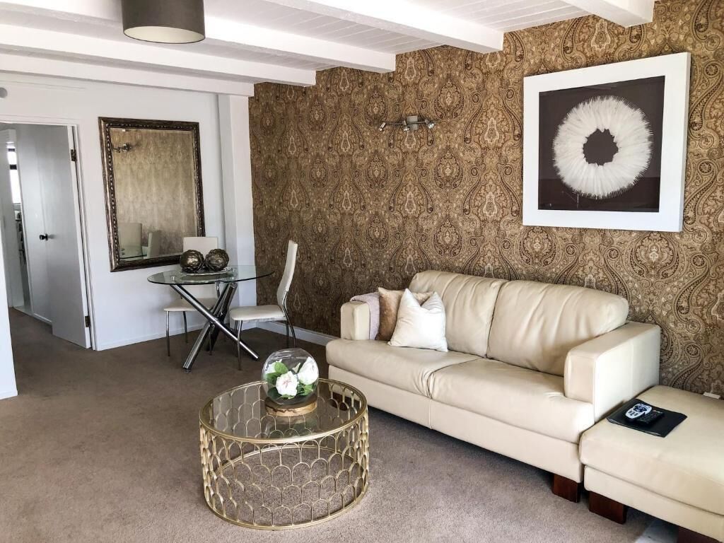 Un salon avec un canapé beige, une table basse en verre, un papier peint à motifs, un tableau encadré et une porte donnant sur une autre pièce.