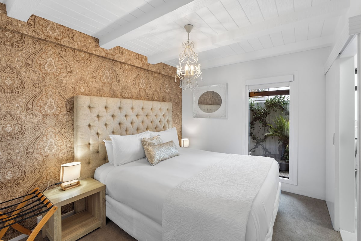 Ein Schlafzimmer mit einer karottenbesetzten Kopfteile, einem Nachttisch mit Lampe, einem großen Bett mit weißer Bettwäsche, einem Chandelier und Blick auf Grün hinter einem Fenster.