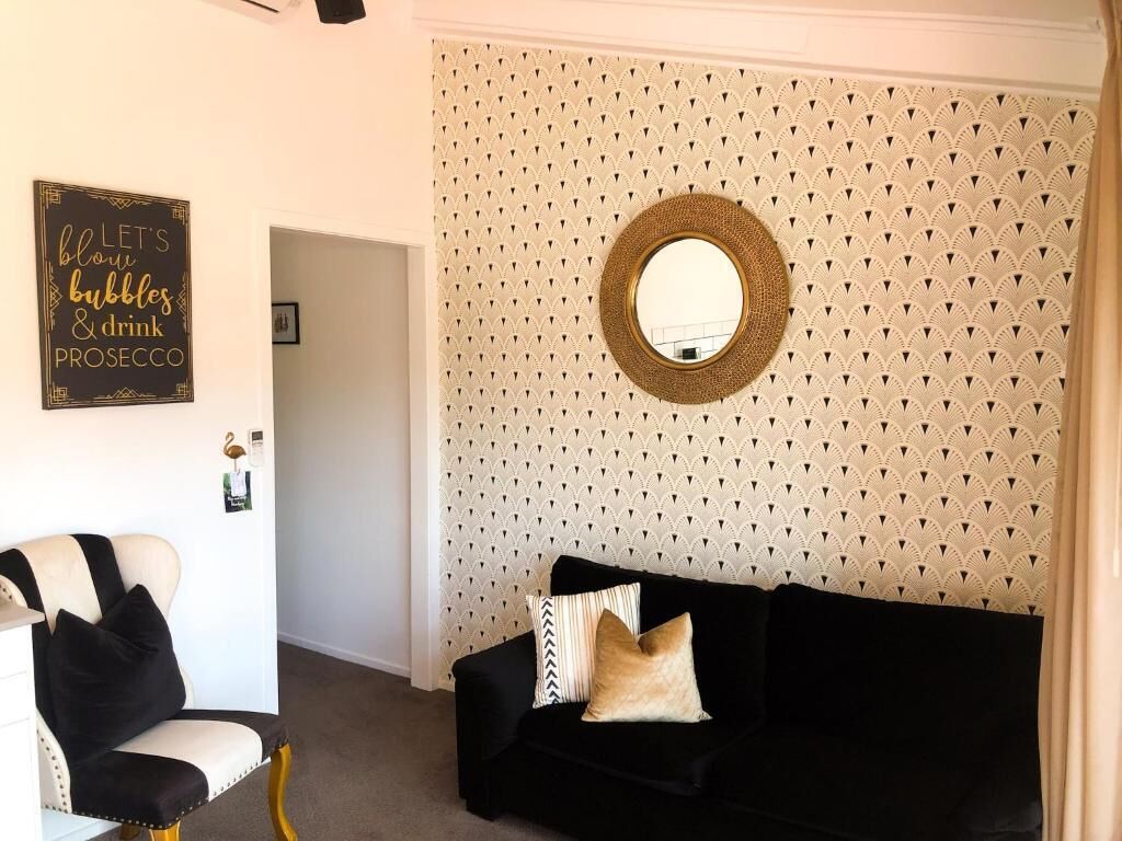Una habitación con papel tapiz a cuadros en blanco y negro, un espejo redondo en la pared, un sofá negro con cojines, y una silla blanca con cojines negros.
