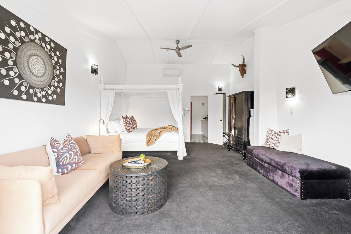 Ein Schlafzimmer mit Baldachinbett, einer Couch, einem Zweisitzer und einem runden Couchtisch sowie einem großen Spiegel.