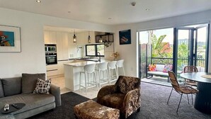 A modern living room with a grey sofa, patterned ottoman, and a dining area with a round table and chairs. A kitchen with white cabinets and a central island with bar stools is visible. - Orewa Beachcomber  (Orewa)