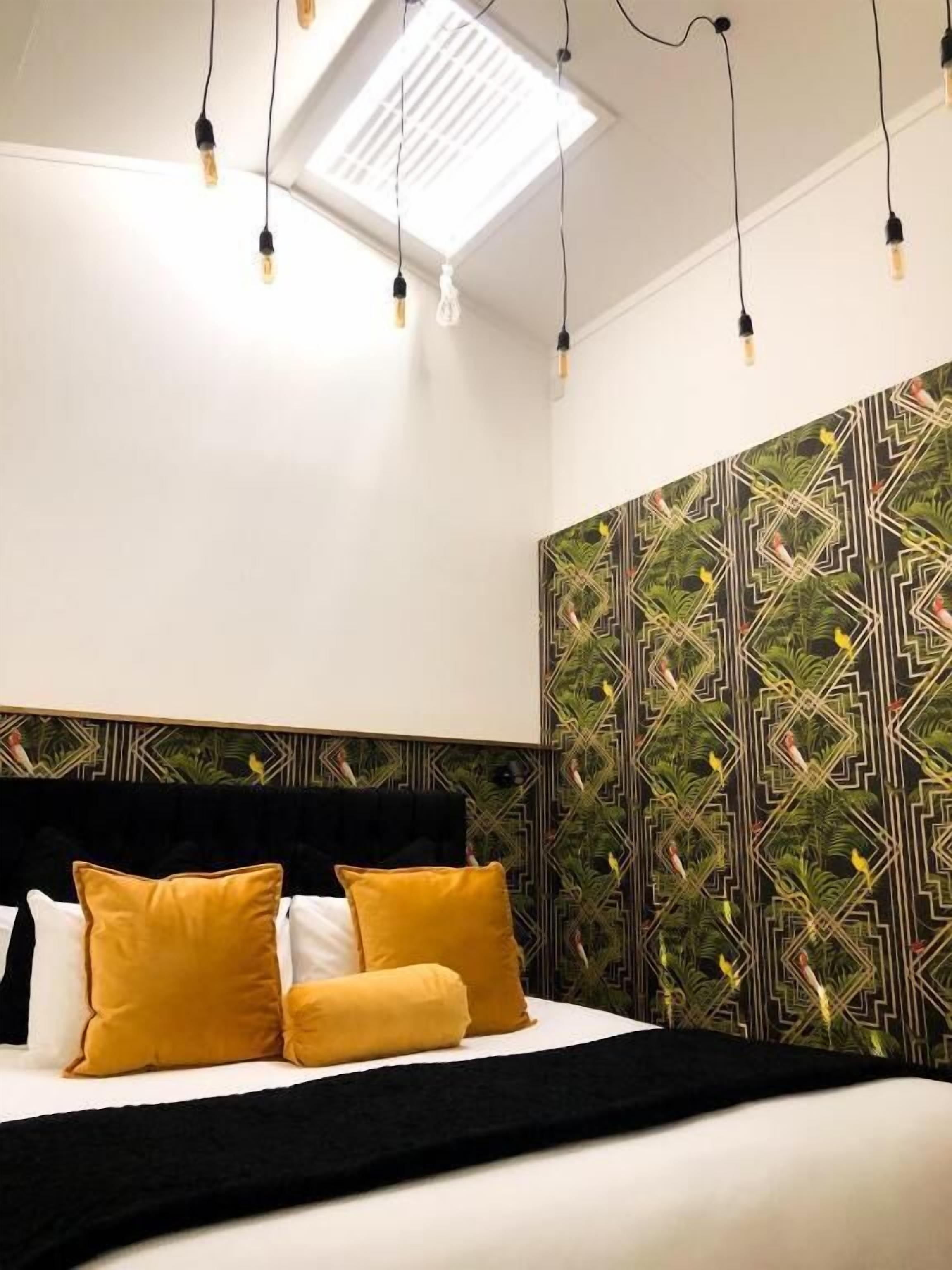 A modern bedroom with a bed, black and yellow pillows, a geometric patterned wall, and a skylight with hanging lights.