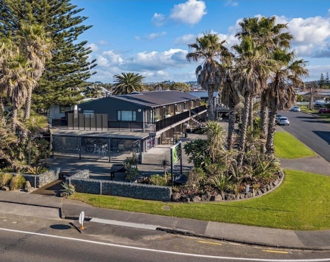 A modern hotel with a flat roof, a covered outdoor seating area, and a landscaped garden with palm trees.