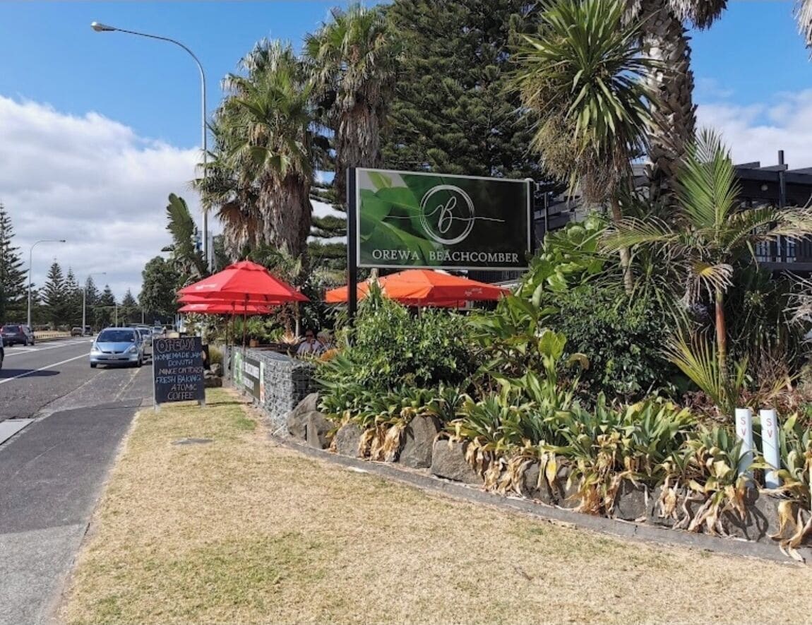 A sign for OREWA BEACHCOMBER with a logo, surrounded by tropical plants and red umbrellas.